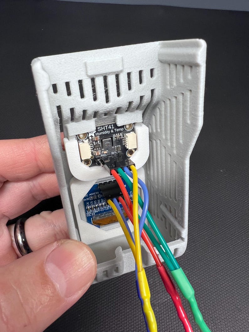 Puede incluir: Una carcasa gris impresa en 3D que contiene una placa de circuito electr&oacute;nico con el texto "SHT41 Humidity & Temp". La placa est&aacute; conectada a cables multicolores, incluyendo rojo, amarillo, verde y azul. El dispositivo se sostiene en una mano.