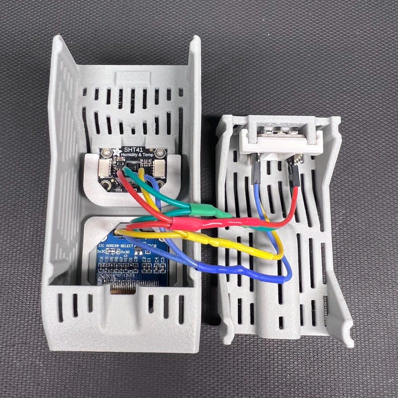 Puede incluir: Dos carcasas grises de componentes electr&oacute;nicos impresas en 3D. Una contiene una placa de circuito etiquetada como "SHT41 Humidity & Temp" y otra con la etiqueta "I2C ADDRESS SELECT". Cables multicolores conectan los componentes.