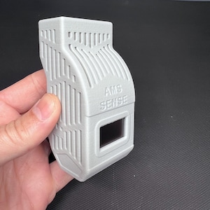 Puede incluir: Un dispositivo rectangular gris claro con el texto "AMS SENSE" y una peque&ntilde;a ventana rectangular. El dispositivo tiene una superficie texturizada con l&iacute;neas verticales y curvas. El dispositivo se sostiene en una mano.