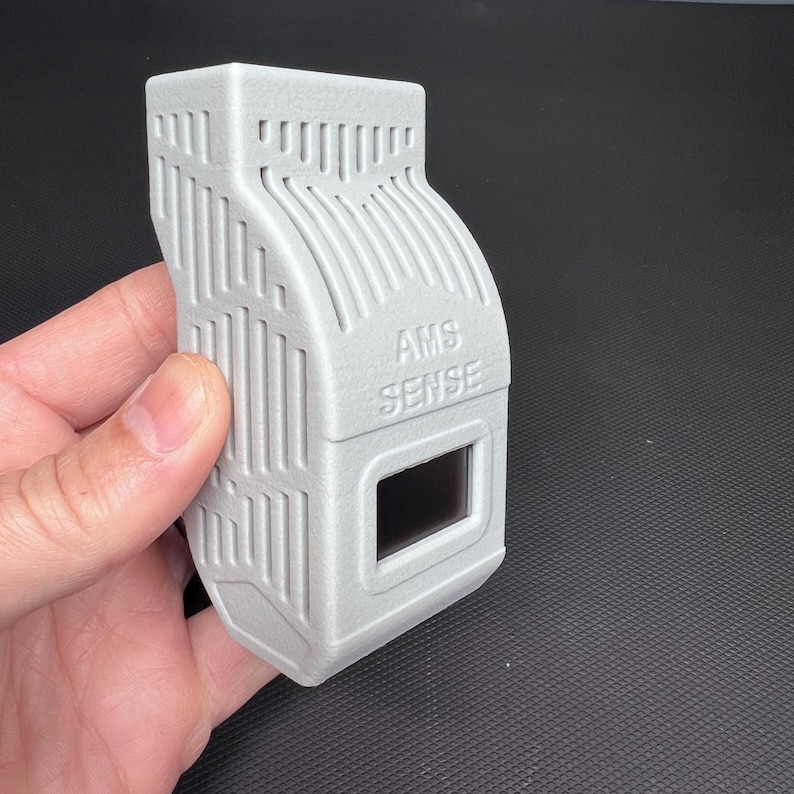 Puede incluir: Un dispositivo rectangular gris claro con el texto "AMS SENSE" y una peque&ntilde;a ventana rectangular. El dispositivo tiene una superficie texturizada con l&iacute;neas verticales y curvas. El dispositivo se sostiene en una mano.