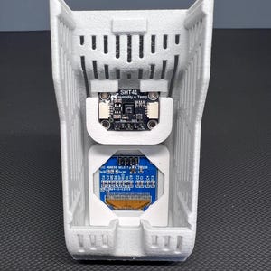 Puede incluir: Una carcasa gris claro, impresa en 3D, con forma rectangular, que alberga dos placas de circuito. La placa superior est&aacute; etiquetada como "SHT41 Humidity & Temp". La placa inferior es azul con varios componentes electr&oacute;nicos y texto. La carcasa tiene un dise&ntilde;o ranurado.