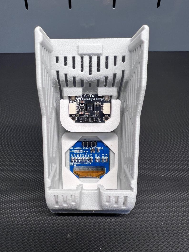 Puede incluir: Una carcasa gris claro, impresa en 3D, con forma rectangular, que alberga dos placas de circuito. La placa superior est&aacute; etiquetada como "SHT41 Humidity & Temp". La placa inferior es azul con varios componentes electr&oacute;nicos y texto. La carcasa tiene un dise&ntilde;o ranurado.