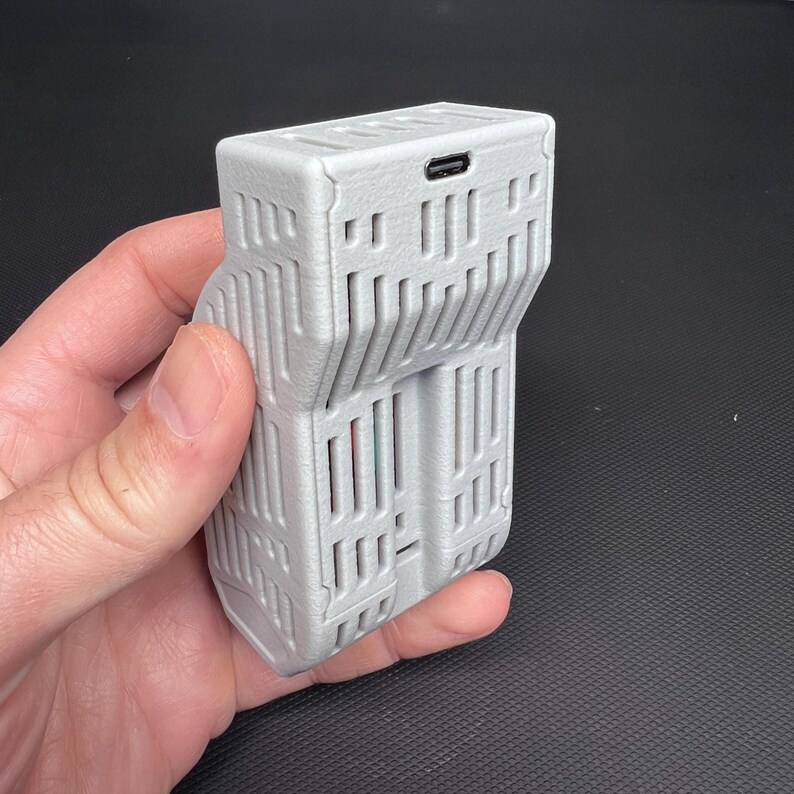 Puede incluir: Un dispositivo rectangular gris claro con una secci&oacute;n articulada. El dispositivo tiene una serie de l&iacute;neas verticales y horizontales, creando un patr&oacute;n similar a una rejilla. Un peque&ntilde;o puerto es visible en la parte superior. El dispositivo se sostiene en una mano.