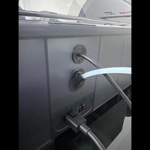 Puede incluir: Primer plano de un dispositivo electr&oacute;nico gris oscuro con un cable de alimentaci&oacute;n negro y un tubo azul claro que se extiende desde la parte trasera. El dispositivo tiene dos ojales de goma negros para la gesti&oacute;n de cables.