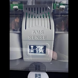 Puede incluir: Un dispositivo gris AMS SENSE con una pantalla digital que muestra la temperatura y la humedad. La pantalla muestra T 75F y H 19%. El dispositivo tiene una parte superior ranurada y est&aacute; encerrado en una carcasa transparente.