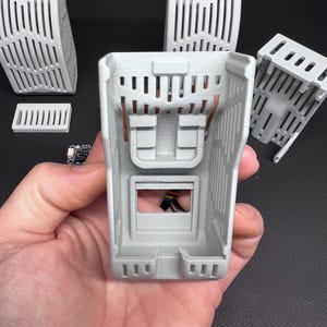 Puede incluir: Un conjunto de componentes grises claros impresos en 3D, que incluyen varias carcasas rectangulares con lados ranurados verticales y una peque&ntilde;a placa de circuito. El componente principal se sostiene en la mano, mostrando su estructura interna y cableado de cobre.