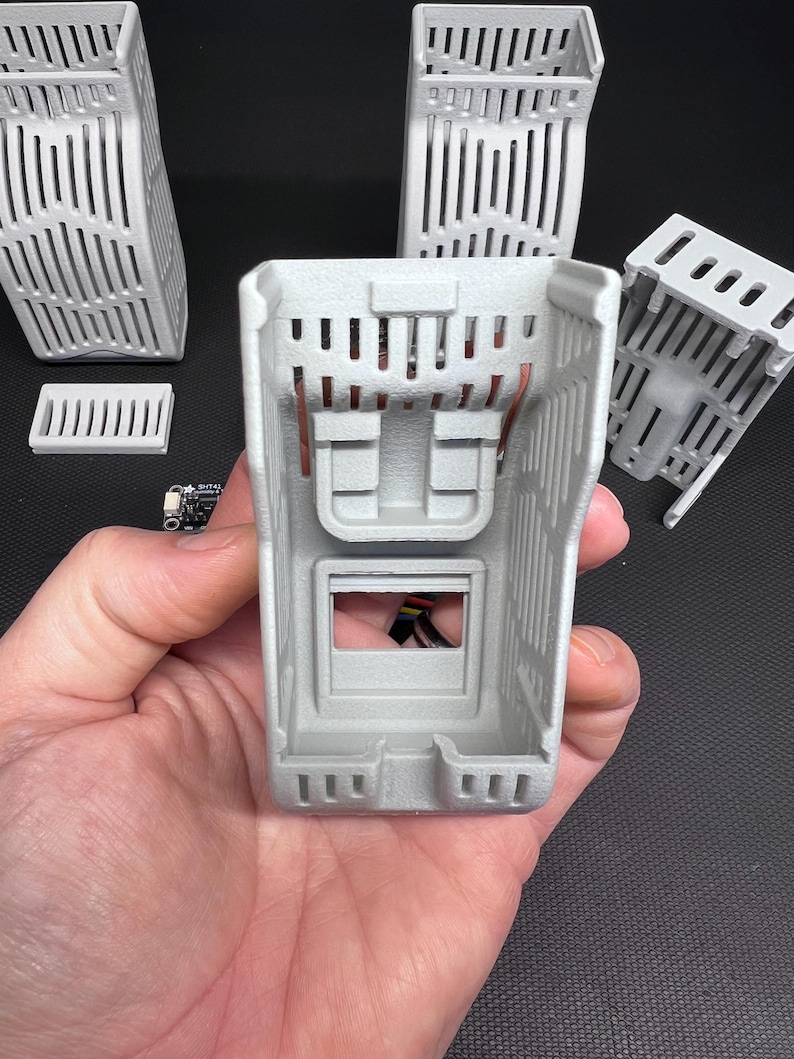 Puede incluir: Un conjunto de componentes grises claros impresos en 3D, que incluyen varias carcasas rectangulares con lados ranurados verticales y una peque&ntilde;a placa de circuito. El componente principal se sostiene en la mano, mostrando su estructura interna y cableado de cobre.