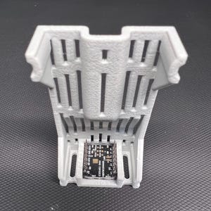 Puede incluir: Un soporte de componentes electr&oacute;nicos gris, impreso en 3D, con una base rectangular y un dise&ntilde;o ranurado. El soporte contiene una peque&ntilde;a placa de circuito con texto y varios componentes. El dise&ntilde;o general es funcional y moderno.