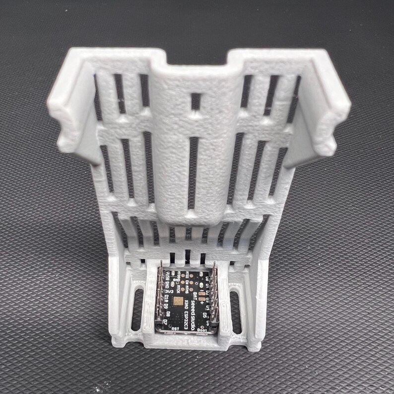 Puede incluir: Un soporte de componentes electr&oacute;nicos gris, impreso en 3D, con una base rectangular y un dise&ntilde;o ranurado. El soporte contiene una peque&ntilde;a placa de circuito con texto y varios componentes. El dise&ntilde;o general es funcional y moderno.