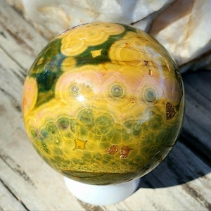 May include: A polished orb of jasper stone, showcasing a blend of yellow, green, and pink hues. The sphere rests on a white base, with intricate patterns and textures visible across its surface. The stone's design includes circular and geometric shapes.
