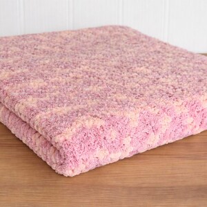 May include: A folded, plush blanket in shades of pink and cream. The blanket has a textured, nubby surface and is resting on a wooden surface. The blanket appears soft and cozy.