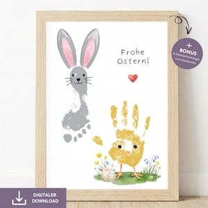 Op de afbeelding: Een ingelijste paasprint met een grijze konijnenvoetafdruk en een gele handafdruk die is getransformeerd tot een kuiken. De print bevat de tekst "Frohe Ostern!" en een rood hart. De lijst is lichtbruin.