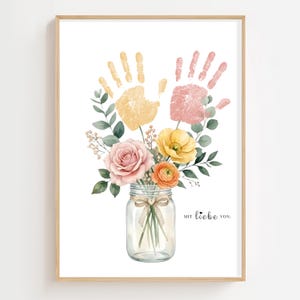 Könnte beinhalten: Gerahmter Aquarelldruck mit einem Blumenstrauß in einem Einmachglas und Handabdrücken darüber. Die Blumen umfassen rosa Rosen, gelbe Mohnblumen und orangefarbene Ranunkeln. Der Text "mit liebe von:" ist aufgedruckt.
