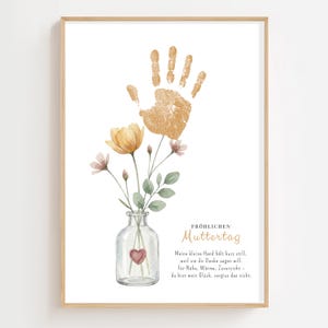 Peut inclure: Une impression encadrée avec une empreinte de main dorée et une illustration à l'aquarelle de fleurs dans un vase en verre. L'impression comprend le texte "FRÖHLICHEN Muttertag" et un poème allemand. L'œuvre est dans un cadre en bois clair.