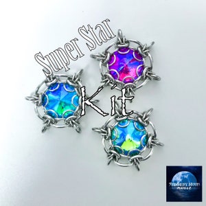 May include: Three silver star-shaped jewelry pieces with iridescent gemstones in blue, green, and purple. The text "Super Star Kit" is written in white on a black background.