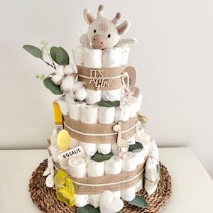 Könnte beinhalten: Eine dreistöckige Wickel-Torte, verziert mit Juteband, Eukalyptusblättern und Wattebäuschen. Die oberste Etage ist mit einem gehäkelten Giraffenspielzeug und einem Holzschild mit der Aufschrift "It's a Girl" geschmückt. Die unterste Etage hat ein Holzschild mit der Aufschrift "Rosalie".