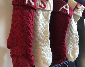 Op de afbeelding: Vier kerstsokken van kabelbreisel hangen op een rij, afwisselend in de kleuren dieprood en crèmewit. Elke sok heeft een geribbelde boord met een enkele geborduurde letter, waarbij de tekens K, S, A en F in contrasterend garen zijn weergegeven.