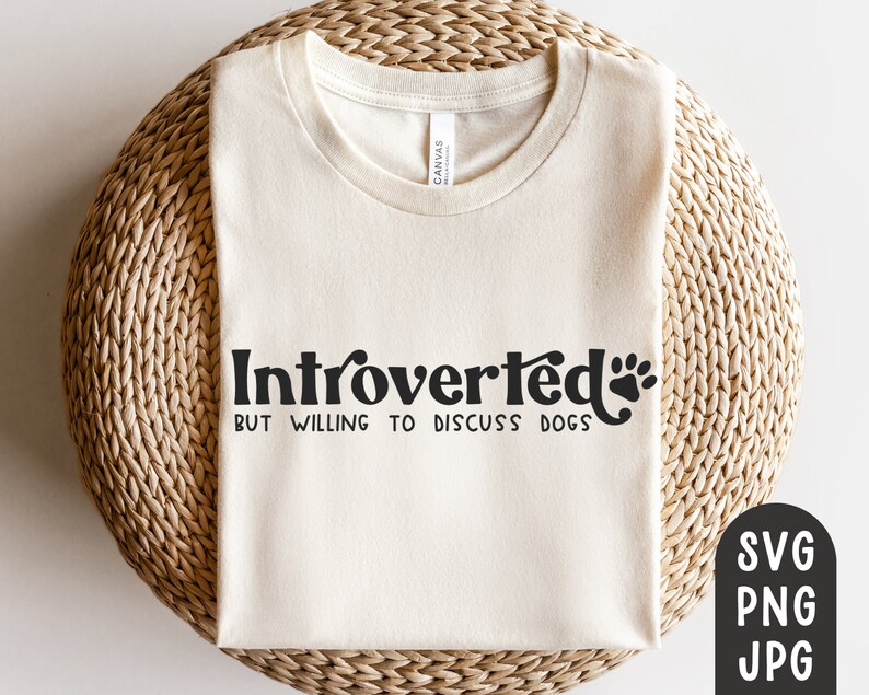 May include: Cream-colored t-shirt with the text "Introverted but willing to discuss dogs" in black. The design includes a paw print. The shirt is folded on a woven, circular surface.