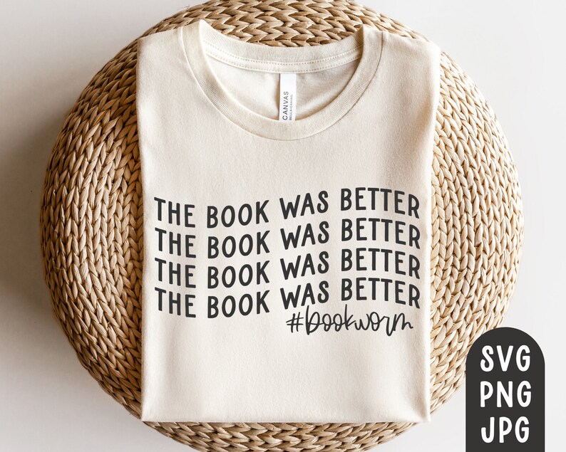 May include: A cream-coloured t-shirt featuring the repeated phrase "THE BOOK WAS BETTER" in black, with the hashtag "#bookworm" underneath. The shirt is presented on a woven, circular mat.