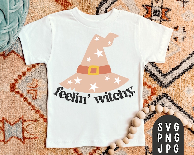 May include: White t-shirt featuring a witch hat design with stars, a brown band, and a gold buckle. The text "feelin' witchy" is printed below the hat in black. The shirt is a children's size.
