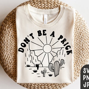 May include: Cream-colored t-shirt with a black graphic design featuring the text "DON'T BE A PRICK" above a desert landscape with a sun, mountains, and cacti. The shirt is made of a soft material and is suitable for casual wear.