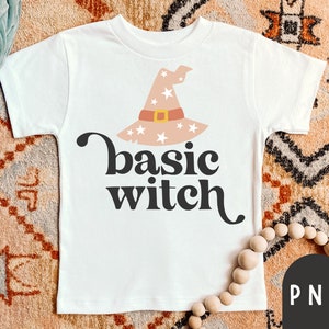 Può includere: T-shirt bianca con grafica di cappello da strega rosa chiaro con stelle e cintura marrone. La scritta "basic witch" è in nero. Un design divertente per Halloween.