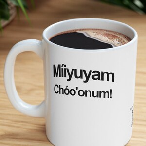 Könnte beinhalten: Eine weiße Keramik-Kaffeetasse mit schwarzem Henkel. Die Tasse hat einen schwarzen Schriftzug mit der Aufschrift "Miíyuyaam Chóo'onum!"