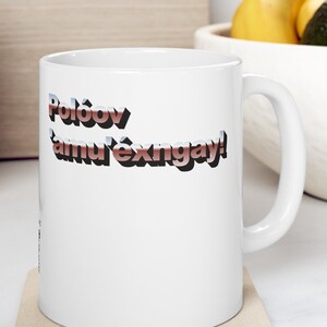 Könnte beinhalten: Weiße Keramiktasse mit einem roten und blauen 3D-Textdesign, das "Poloov tamu'exngay!" lautet.