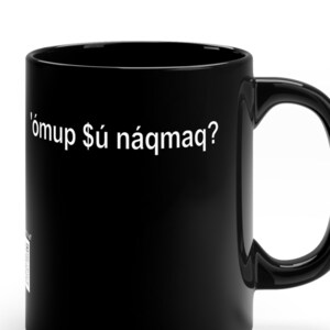 Könnte beinhalten: Schwarze Keramik-Tasse mit gebogenem Henkel. Die Tasse zeigt den weißen Text "'ómuṕ $ú náqmaq?" auf der Vorderseite. Die Tasse hat eine Standardgröße und -form, geeignet für heiße oder kalte Getränke.