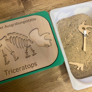 Könnte beinhalten: Ein hölzernes Dinosaurier-Grabungsset mit grünem Rand. Das Set enthält eine Holzschale mit einem Triceratops-Skelett-Ausschnitt und einen separaten Behälter gefüllt mit Sand. Der Sand enthält Holzstücke, die ausgegraben werden können, um das Skelett zusammenzubauen.