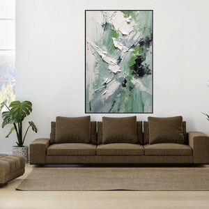 Könnte beinhalten: Ein braunes Sofa mit drei Kissen steht vor einer weißen Wand mit einem gerahmten abstrakten Gemälde mit grünen, weißen und schwarzen Farbschlägen. Links vom Sofa steht eine Topfpflanze und rechts vom Sofa eine weitere Topfpflanze.