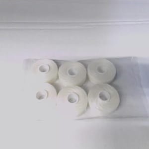 May include: Six white spools of thread are arranged in a clear plastic bag. The spools are round with a central hole, and the thread is tightly wound. The background is a plain white surface.