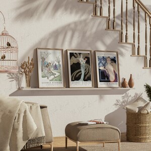 May include: Three framed art prints with a white background and gold frames. The prints feature illustrations of women in vintage clothing. The prints are titled "George Barbier" and are displayed on a shelf with a birdcage, a vase of wheat, and two decorative vases.