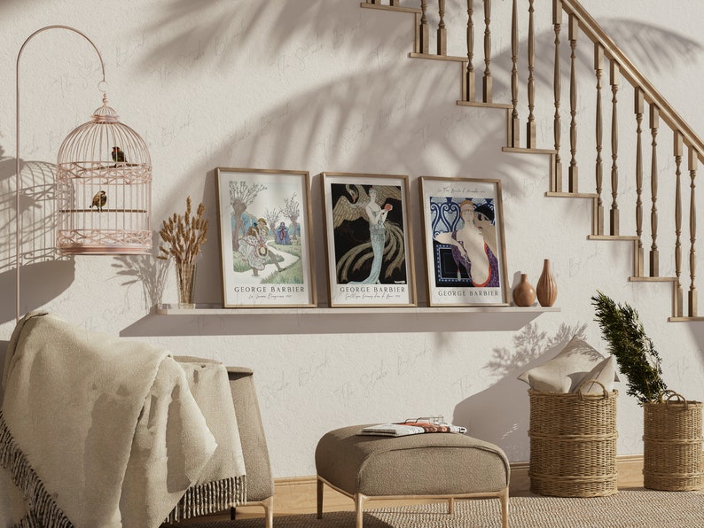May include: Three framed art prints with a white background and gold frames. The prints feature illustrations of women in vintage clothing. The prints are titled "George Barbier" and are displayed on a shelf with a birdcage, a vase of wheat, and two decorative vases.