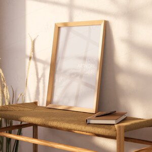Puede incluir: Un marco de fotos de madera con un lienzo blanco en blanco, sentado en un banco de madera con una libreta de cuero marrón y un bolígrafo. El fondo es una pared beige claro con sombras de una ventana.