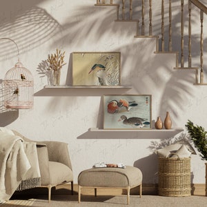 May include: A living room interior with a beige armchair, a throw blanket, a footstool, and two framed bird illustrations on the wall. There are two wicker baskets, a birdcage, and a vase with dried flowers on a shelf.