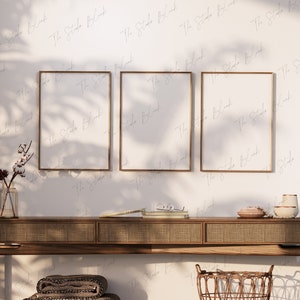 May include: Three gold framed empty picture frames hang above a wooden console table with a woven rattan top. The console table has a woven rattan basket underneath. The wall behind the table is white with a subtle pattern of light gray script.