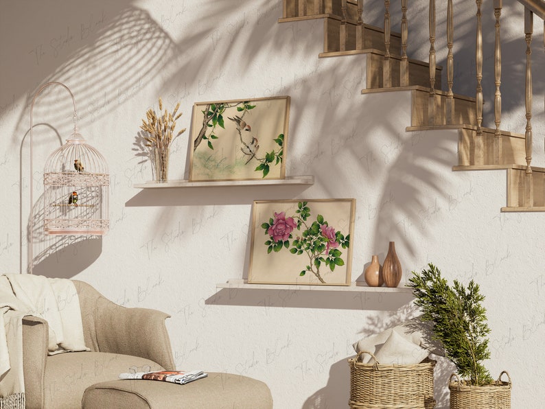 May include: Two framed botanical prints, one with pink flowers and the other with birds on branches, are displayed on shelves against a white wall. A birdcage with a pink finish and small birds inside sits on a beige armchair. A potted green plant and two brown vases are on the shelf below the prints.