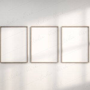May include: Three empty picture frames with a light wood finish, hanging on a white wall. The frames are arranged in a row, with the middle frame slightly higher than the other two.