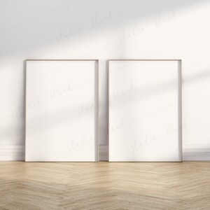 May include: Two empty picture frames with thin gold frames are hanging on a white wall. The frames are positioned side by side, with a wooden floor in the foreground.