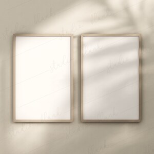 Könnte beinhalten: Zwei leere Bilderrahmen mit einem hellen Holzrahmen, die an einer hellbeigen Wand hängen. Die Rahmen sind nebeneinander angeordnet, mit einem kleinen Abstand zwischen ihnen. Die Wand wird von natürlichem Licht beleuchtet, das weiche Schatten auf die Rahmen wirft.