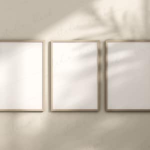 May include: Three blank frames with a light brown wood finish are hanging on a beige wall. The frames are arranged in a row, with the middle frame slightly higher than the other two. The frames are casting shadows on the wall, creating a minimalist and modern aesthetic.