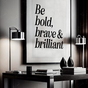 May include: Black and white motivational poster with the text "Be bold, brave & brilliant" hanging on a wall above a black desk with two lamps and a stack of books.