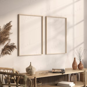 May include: Two empty picture frames with light wood frames hang on a white wall. The frames are side by side and are both vertical. There is a wooden table with a vase and books on it in front of the frames.
