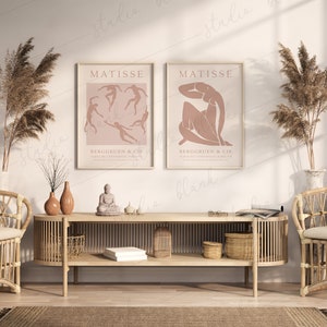 May include: Two framed art prints with a pink background and black line drawings of people. The prints are titled "MATISSE" and have the text "BERGGREEN & CIE" below the drawings. The prints are hanging on a white wall above a light brown wooden shelf with a small Buddha statue and other decorative items.