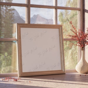 May include: A gold-toned picture frame with a white mat and a blank white canvas inside. The frame is sitting on a wooden surface next to a vase with red berries and a smaller vase.