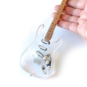Puede incluir: Una guitarra eléctrica de acrílico transparente con un mástil de madera natural y herrajes cromados. La guitarra es una réplica en miniatura de una Fender Stratocaster.