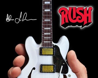 Op de afbeelding: Witte elektrische gitaar met gouden hardware en een zwarte pickguard. De gitaar heeft een Gibson-logo op de body en het woord "Rush" in rode letters staat op de achtergrond.