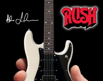 Op de afbeelding: Een witte elektrische gitaar met een zwarte pickguard en een zwart-wit gestreept toets. De gitaar wordt in een hand gehouden. De handtekening "Alex Lifeson" is zichtbaar in de linkerbovenhoek van de afbeelding. Het woord "RUSH" is zichtbaar in de rechterbovenhoek van de afbeelding.