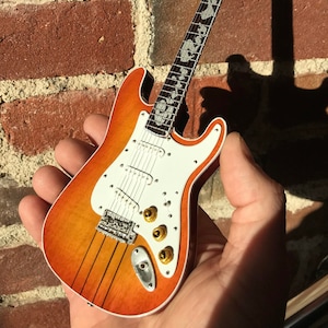 May include: A miniature electric guitar with a sunburst finish and white pickguard. The guitar has a silver-colored tuning pegs and a white inlay on the fretboard.